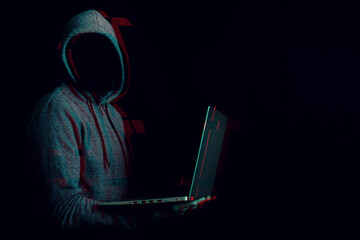 Man without a face in a hood holds a laptop in his hands on a dark background. Concept of cyber...