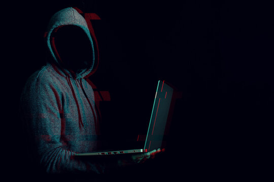 Man without a face in a hood holds a laptop in his hands on a dark background. Concept of cyber rogue, hacker. Banner. Added glitch effect