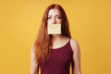 young woman hiding sadness or depression behind fake smile drawn on yellow sticky note paper