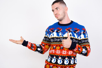 Happy cheerful Young handsome Caucasian man wearing Christmas sweater against white wall showing thumb up and pointing with the other hand. I recommend this.