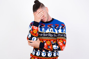Young handsome Caucasian man wearing Christmas sweater against white wall making facepalm gesture while smiling amazed with stupid situation.