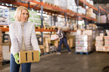 Portrait of tired middle aged woman working in large warehouse, carrying boxes