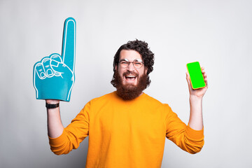 Excited young bearded man is holding a blue fan glove and a phone laughing.