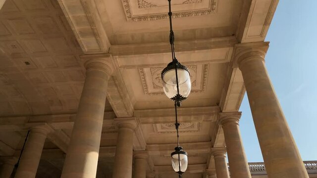 Footage of hung lamps and columns at a palace called "Domaine National du Palais Royal" in Paris. It is a sunny summer day. Camera moves forward. Sun rays effects occur.