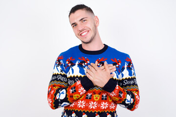 Honest Young handsome Caucasian man wearing Christmas sweater against white wall keeps hands on...