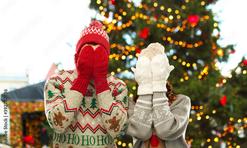 Poster winter holidays and people concept - couple in sweaters, hats and woolen mittens covering their faces over christmas tree lights on background