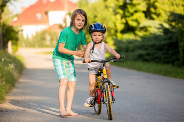 Fototapeta premium Young mother, helpOlder brother, helping his little brother to learn how to ride a bike, holding him and teaching him bikinging her son to learn how to ride a bike, holding him and teaching him