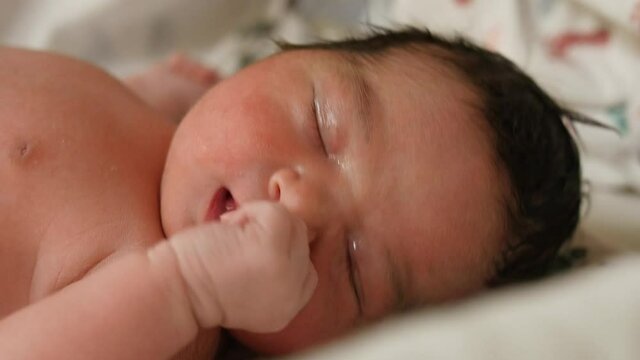 Close-up of newborn baby soon after her birth. She lies on a clean bed quietly and restlessly, coming in and out of her sleep.  The antibiotic ointment applied on her eyes is still visible.