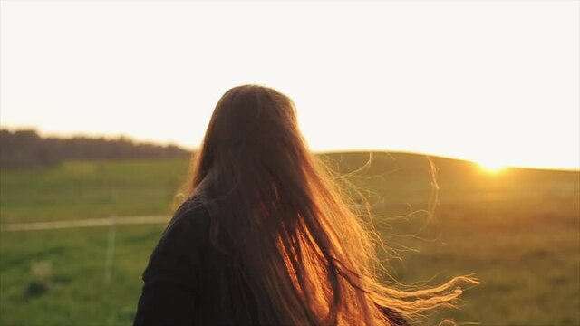 An enthusiastic long-haired girl spins and jumps against the backdrop of a sunset on a green field. Slow motion