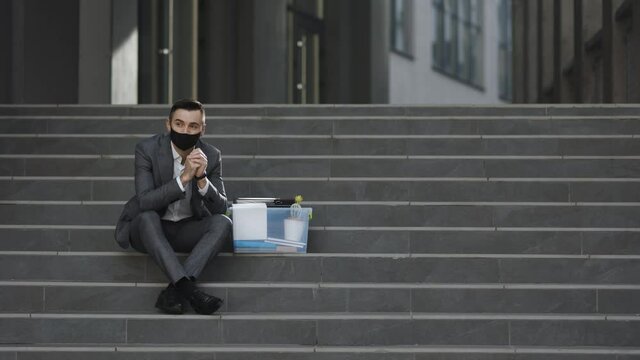 Unemployed businessman lost his business. Fired male office worker in medical mask sitting on stairs in depression with box of stuff. Anxious concept. Workless man in despair
