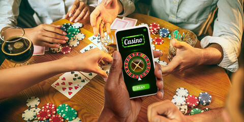 Online gambling, casino concept. Hand holding device with lottery, casino cover. Playing dips and cards on table on the background. Poker, bookmaking, gaming, modern technologies, business and finance
