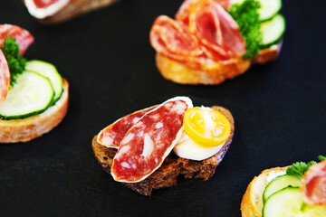 Sandwiches with smoked sausage, decorated with cucumbers and yellow tomatoes on a black background