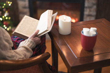 Fototapeta na wymiar Woman hand with old retro book and cup of hot cocoa and marshmallow on wooden table near christmas tree and fireplace. Toned