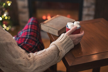Obraz na płótnie Canvas Woman hand with old retro book and cup of hot cocoa and marshmallow on wooden table near christmas tree and fireplace. Toned