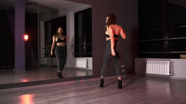 Young energetic woman in high heels dancing modern dance, performs plastic movements in a studio with large mirrors. Slow motion.