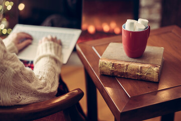 Woman hand with laptop, cup of hot cocoa and marshmallow on book on wooden table near christmas tree and fireplace. Toned