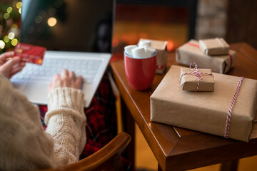 Woman holding credit card using laptop for making order sitting at table with packaging gift, cup of cocoa and marshmallows near fireplace and christmas tree. Online shopping concept