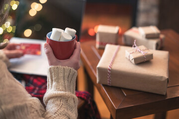 Fototapeta na wymiar Woman holding credit card using laptop for making order sitting at table with packaging gift, cup of cocoa and marshmallows near fireplace and christmas tree. Online shopping concept