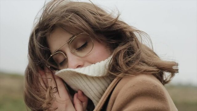 Portrait of a sensual young woman standing against the sky and pulling the collar of a sweater over her nose with closed eyes
