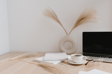 Female home office desk workspace. Blank screen laptop computer with copy space. Coffee cup, pampas grass in stylish vase, stationery on beige wooden table. Minimalist lifestyle blog mockup.