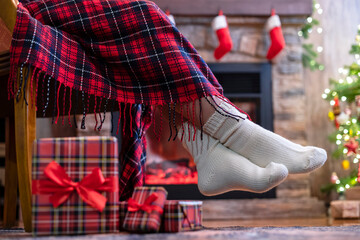 Fototapeta na wymiar Woman legs in a winter socks covered plaid sitting and relaxation on armchair near fireplace and christmas tree pakking gift boxes for family. Bottom view.