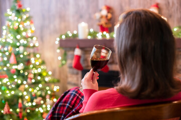 Back view of woman drinking wine from glass covered plaid sitting and relaxation on armchair near christmas fareplace and fir-tree.
