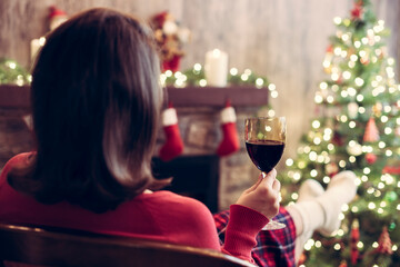 Back view of woman drinking wine from glass covered plaid sitting and relaxation on armchair near christmas fareplace and fir-tree.