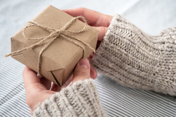 A girl in a sweater holds a gift decorated with her own hands. Beautiful gift box made of Kraft paper for birthday, Christmas or new year. Gift in women's hands, close-up. Holiday background