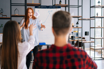 Redhead young business woman finishing report on white board. Dissatisfied colleagues throw crumpled paper balls at frightened lady. Confused female runs away and hides behind blackboard.