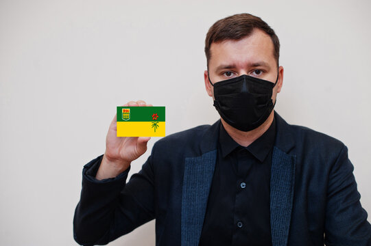 Man wear black formal and protect face mask, hold Saskatchewan flag card isolated on white background. Canada provinces coronavirus Covid country concept.