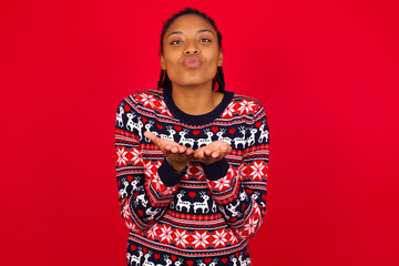 Young beautiful African American woman standing against red background sending blow kiss with pout lips and holding palms to send air kiss.