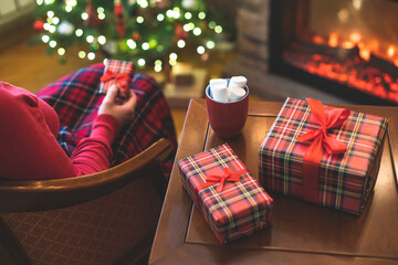 Alone woman covered plaid with cup of cocoa with marshmallows sitting and relaxation on armchair near fareplace and christmas tree after finishing pakking gift boxes for fami