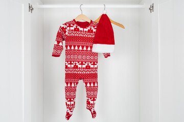 White and red christmas hat and pajamas hanged on a wooden hangers in the middle of a white closet.