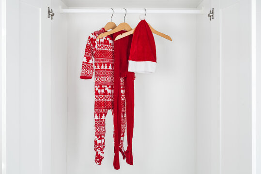 White and red christmas pajamas, hat and tights hanging on a wooden hangers in the middle of a white closet.