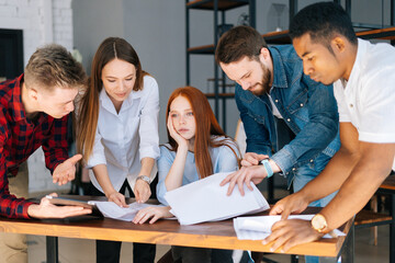Exhausted tired young business woman sitting at desk, colleagues shaking papers in face paper documents. Businesswoman under stress at workplace. Female boss exhausted by management work.