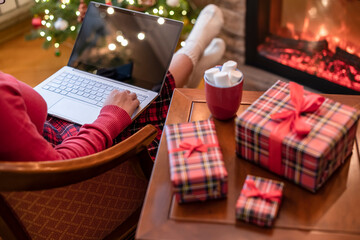 Naklejka na ściany i meble Christmas. Woman using laptop for searching gift ideas sitting at table with ready gift boxes and cup of cocoa and marshmallows near fireplace and christmas tree. Concept