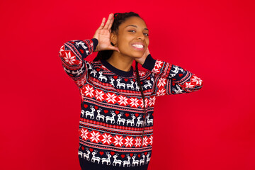 Young beautiful African American woman standing against red background Trying to hear both hands on ear gesture, curious for gossip. Hearing problem, deaf