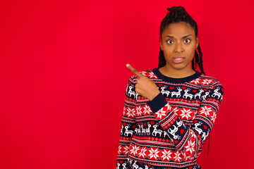 Young beautiful African American woman standing against red background Pointing aside worried and...
