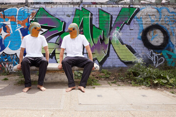 Two men in latex bird masks talk to each other on a seat in front of a graffitied wall.  It is urban.
