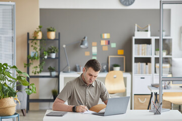 Young businessman sitting at the table in front of the laptop and writing plans in his note pad he working at office © AnnaStills