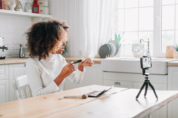 Serious trendy kinky haired blogger making bronzer swatches on hand using makeup brush against smartphone on tripod closeup