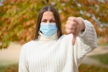 Front view portrait of serious girl doing thumbs down gesture with protective mask on city street