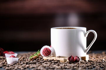 White cup full of coffee beans , red flowers and red chocolate candy against the background of a red wood wall. Morning espresso. Coffee mug.	