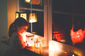 Little kid girl sitting by window with selfmade hand crafted lanterns with candles for St. Martin...