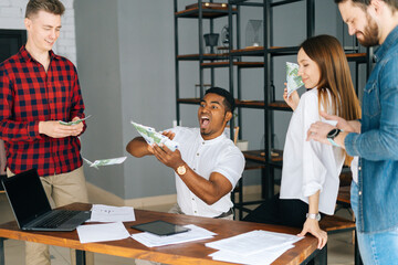 Happy business team of young multi-ethnic employees celebrating victory and big profit at office workplace. Cheerful excited young business man and woman throwing away banknote. Rain of money.