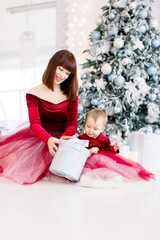 Close up studio portrait of happy mother and little daughter in beautiful wine red dresses, sitting near christmas tree, and opening Christmas gift. Happy holidays concept