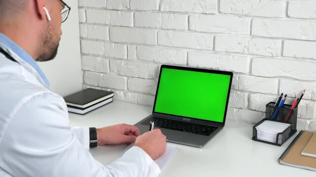 Man doctor wears medical coat wireless earphones sits on chair at desk in hospital office looking green screen laptop computer listens patient client writes symptoms in notebook, social distancing