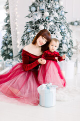 Shot of beautiful mother in fashion red dress posing with her cute little baby girl in cozy light decorated studio. Mother and daughter concept. New Year and Christmas