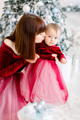 Christmas holidays, family concept. Pretty young mom kissing her little sweet beautiful daughter, spending winter time together, sitting on the floor in front of Christmas tree