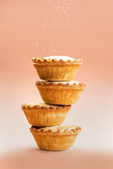 Mince pies stacked on each other on pink background with sugar sifting on top. A mince pie is a traditional Christmas sweet pie, filled with a mixture of dried fruits and spices. - 392237822